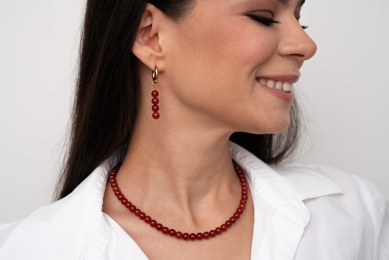 May include: A red carnelian necklace and earrings set. The necklace is made of small, round beads and the earrings are dangle earrings with a gold hoop and red carnelian beads.