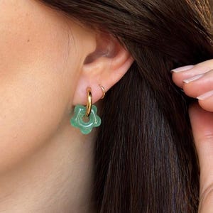 May include: A gold hoop earring featuring a small, light green, five-petaled flower charm made of a translucent, smooth material. The earring is worn in the earlobe, with a simple, thin gold hoop visible in the upper cartilage area.