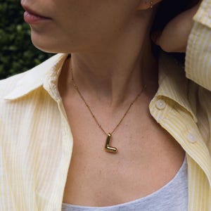 May include: A gold-coloured necklace featuring the letter "L" pendant. The necklace is worn over a grey top and a yellow and white striped button-down shirt. The background is a green bush.