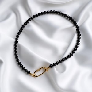 May include: A black beaded necklace with a gold-coloured clasp. The necklace features small, round, black beads. The clasp is an oval shape. The necklace is displayed on white, silky fabric.