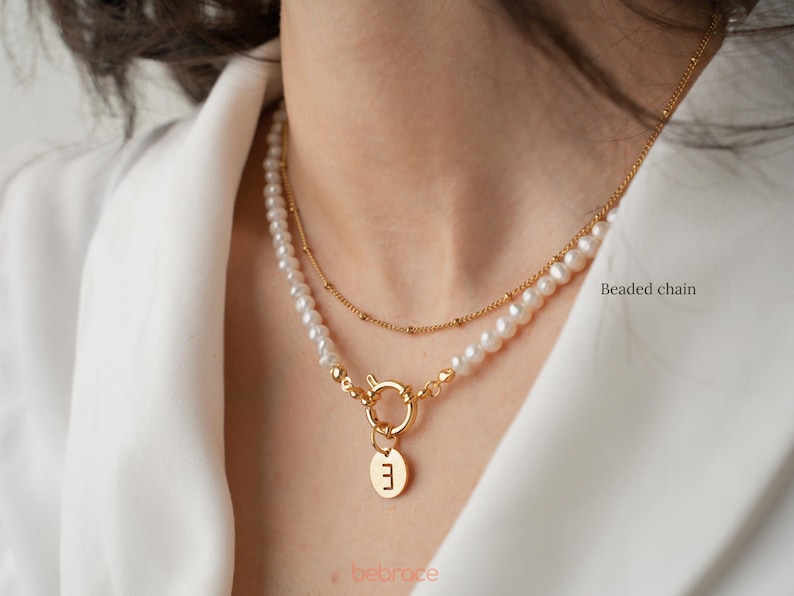 Op de afbeelding: Een gouden ketting met een parelstreng en een gouden bedel met de letter "E" die eraan hangt. De ketting is gelegd over een kralenketting. "Kralenketting" staat in de rechterbenedenhoek van de afbeelding.