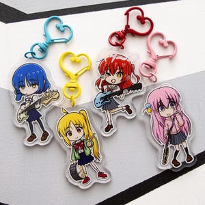 Puede incluir: Una colección de llaveros de estilo anime con personajes coloridos con instrumentos musicales. Cada llavero tiene un cierre en forma de corazón en varios colores, incluyendo azul, rojo y rosa. Los personajes están representados con guitarras y tambores, vistiendo uniformes escolares.