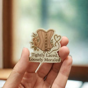 May include: A sticker featuring a light pink corset design with brown lacing and floral accents. The text "Tightly Laced, Loosely Moraled" is printed below the corset. The sticker is held in a hand.