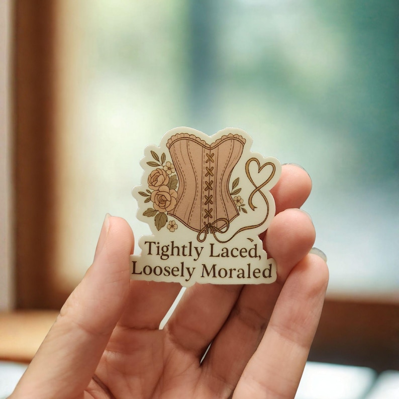 May include: A sticker featuring a light pink corset design with brown lacing and floral accents. The text "Tightly Laced, Loosely Moraled" is printed below the corset. The sticker is held in a hand.
