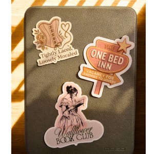 May include: A collection of stickers on a gray surface. The stickers feature a corset with text "Tightly Laced, Loosely Moraled", a sign that reads "The One Bed Inn, Vacancy for Enemies to Lovers", and a woman reading with "Wallflower Book Club".
