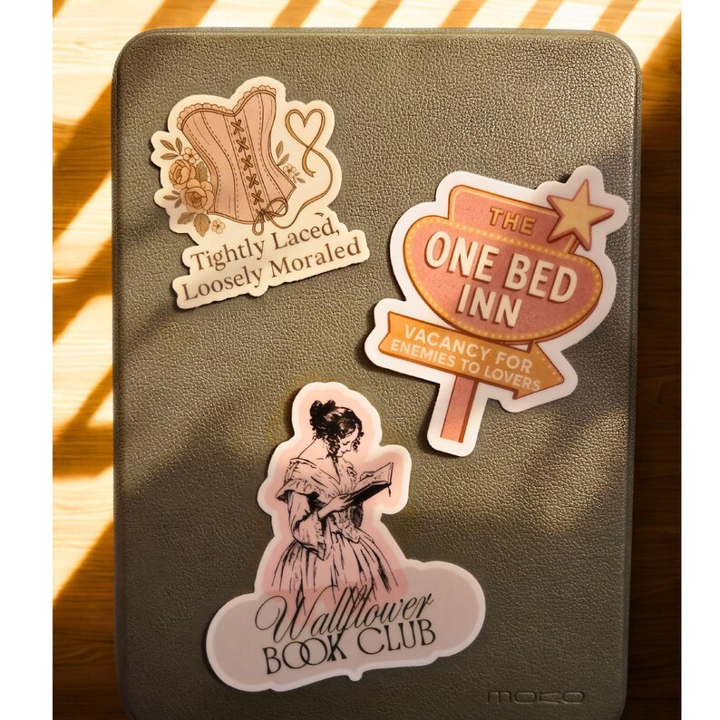 May include: A collection of stickers on a gray surface. The stickers feature a corset with text "Tightly Laced, Loosely Moraled", a sign that reads "The One Bed Inn, Vacancy for Enemies to Lovers", and a woman reading with "Wallflower Book Club".