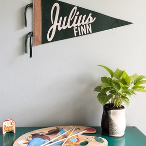 Puede incluir: Un bander&iacute;n de fieltro verde oscuro con el nombre "Julius Finn" en escritura blanca. El bander&iacute;n tiene un borde beige y cintas verdes para colgar. Una paleta de artista de madera con pintura y pinceles est&aacute; sobre una superficie turquesa.