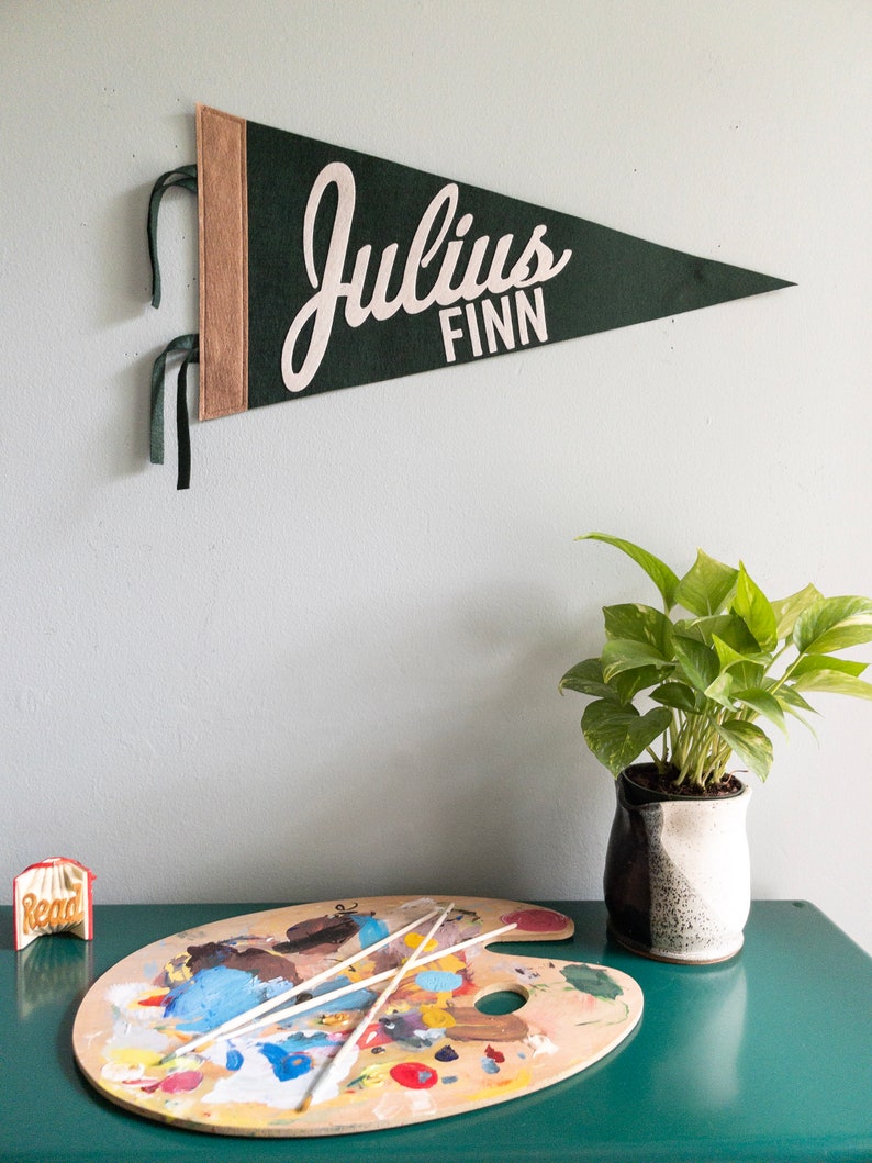 Puede incluir: Un bander&iacute;n de fieltro verde oscuro con el nombre "Julius Finn" en escritura blanca. El bander&iacute;n tiene un borde beige y cintas verdes para colgar. Una paleta de artista de madera con pintura y pinceles est&aacute; sobre una superficie turquesa.