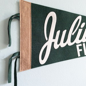 Puede incluir: Un bander&iacute;n de fieltro verde oscuro con el nombre "Julius" en escritura blanca. El bander&iacute;n tiene un borde beige en el lado izquierdo y cintas verdes para colgar. El bander&iacute;n se muestra contra una pared blanca.