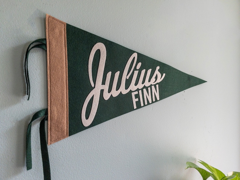 Puede incluir: Un bander&iacute;n de fieltro verde oscuro con el nombre "Julius Finn" en letras beige claro. El bander&iacute;n tiene un borde beige en el lado izquierdo y cintas verdes oscuras para colgarlo. Montado en una pared azul claro.