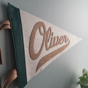 以下が含まれることがあります： 茶色の筆記体で「Oliver」という名前が入った、三角形のフェルトペナント。 ペナントは、薄いベージュの背景、濃い緑色の縁取り、タン色の吊り下げストラップが付いています。 ペナントは水色の壁に掛けられています。