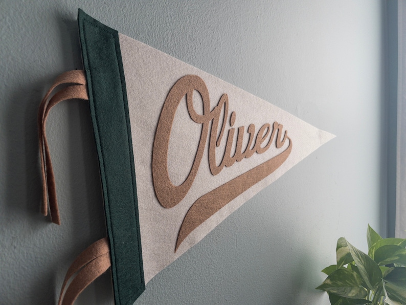 以下が含まれることがあります： 茶色の筆記体で「Oliver」という名前が入った、三角形のフェルトペナント。 ペナントは、薄いベージュの背景、濃い緑色の縁取り、タン色の吊り下げストラップが付いています。 ペナントは水色の壁に掛けられています。