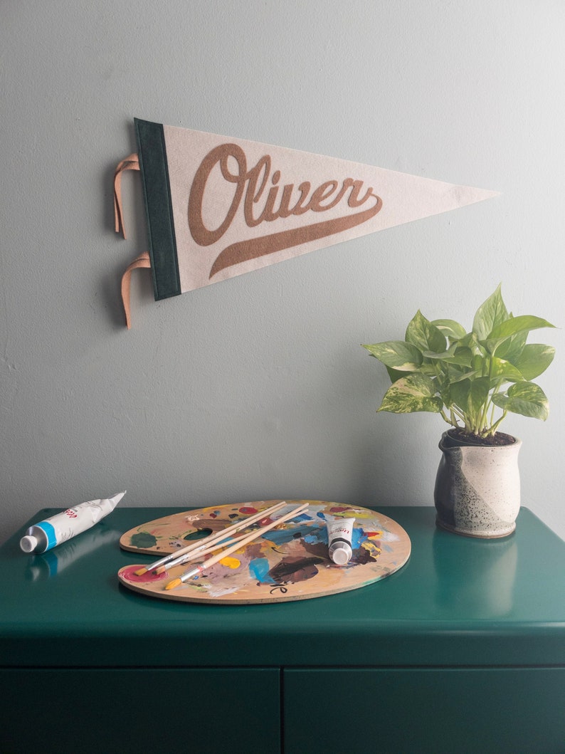 以下が含まれることがあります： 「Oliver」という名前が茶色の筆記体で書かれたベージュ色のペナントが灰色の壁に掛けられています。その下に、絵の具のチューブと筆が置かれた木製のアーティストパレットが、濃い緑色の表面に置かれ、鉢植えの植物の隣にあります。