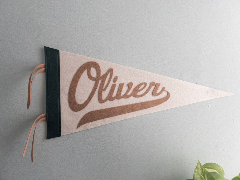以下が含まれることがあります： ベージュのフェルトペナントに、茶色の筆記体で「Oliver」と書かれています。ペナントには濃い緑色の縁取りと、吊り下げ用のタン革の紐が付いています。ペナントは明るい灰色の壁に飾られています。