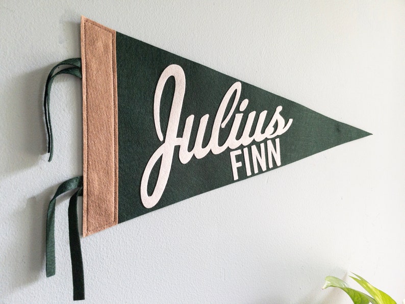 Puede incluir: Un bander&iacute;n de fieltro verde oscuro con el nombre "Julius Finn" en escritura blanca. El bander&iacute;n tiene un borde beige en el lado izquierdo y cintas verdes para colgar. Un art&iacute;culo decorativo para la habitaci&oacute;n de un ni&ntilde;o.