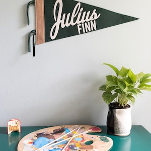 Puede incluir: Un bander&iacute;n de fieltro verde oscuro con el nombre "Julius FINN" en letras blancas cuelga de una pared. El bander&iacute;n tiene un borde beige y cintas verdes. Debajo, una paleta de artista de madera con pintura y pinceles.