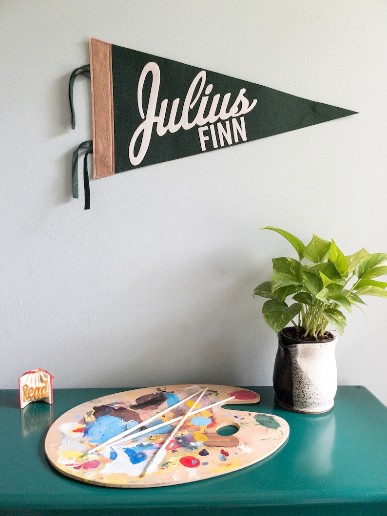 Puede incluir: Un bander&iacute;n de fieltro verde oscuro con el nombre "Julius FINN" en letras blancas cuelga de una pared. El bander&iacute;n tiene un borde beige y cintas verdes. Debajo, una paleta de artista de madera con pintura y pinceles.
