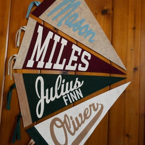 May include: A collection of personalized felt pennants in various colors, including beige, burgundy, green, and white. Each pennant displays a name in a contrasting color, such as blue, white, or brown. The pennants are hanging against a wooden background.