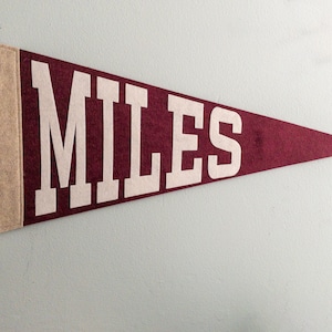 Puede incluir: Bandera de fieltro granate con la palabra "MILES" en letras blancas. La bandera tiene un borde de fieltro beige en el lado izquierdo con lazos beige para colgar. Un art&iacute;culo de recuerdo o decoraci&oacute;n cl&aacute;sico.