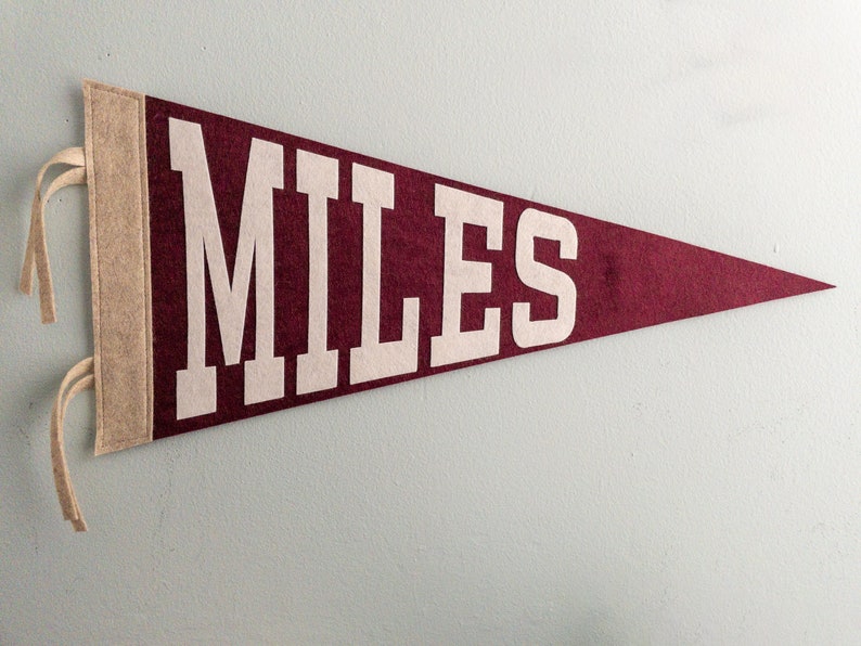 Puede incluir: Bandera de fieltro granate con la palabra "MILES" en letras blancas. La bandera tiene un borde de fieltro beige en el lado izquierdo con lazos beige para colgar. Un art&iacute;culo de recuerdo o decoraci&oacute;n cl&aacute;sico.