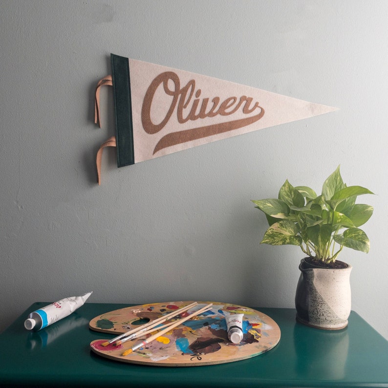 以下が含まれることがあります： グレーの壁に、茶色の筆記体で「Oliver」と書かれたベージュのフェルトペナントが吊り下げられています。ペナントは濃い緑色の縁と革のストラップが付いています。緑色のテーブルには、絵の具と筆の入った木製のアーティストパレットが置かれ、その隣には鉢植えの植物があります。