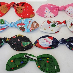 May include: Seven Christmas-themed fabric hair bows with various designs, including Santa Claus, gingerbread men, Christmas trees, and reindeer. The bows are tied with a knot in the center and have a festive holiday feel.