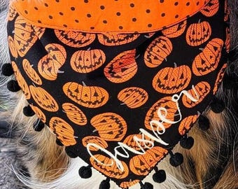 May include: A triangular pet bandana featuring a black fabric base with an orange pumpkin print and a solid orange top border with black polka dots. The bottom edge is trimmed with small black pom-poms and the name Jasper is embroidered in white.