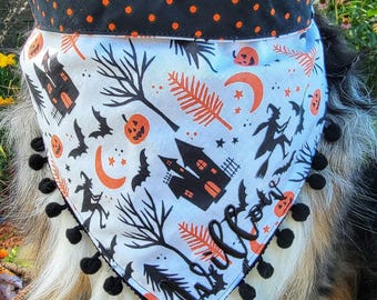 May include: A triangular pet bandana featuring a white background with a black and orange Halloween print of haunted houses, witches, bats, pumpkins, and crescent moons. The top band is black with orange polka dots and black pom-pom trim.
