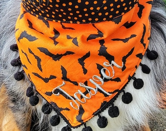 May include: A triangular pet bandana featuring an orange fabric with a black bat pattern and a black collar section with orange polka dots. The bottom edge is trimmed with black pom-poms, and the name Jasper is embroidered in silver thread.