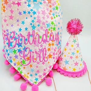 May include: A white birthday bandana and party hat set. The bandana and hat are covered in colorful stars and feature the embroidered text "Birthday Girl". Both items are trimmed with pink pom-poms.