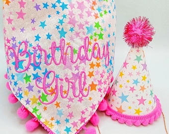 May include: A white birthday bandana and party hat set. The bandana and hat are covered in colorful stars and feature the embroidered text "Birthday Girl". Both items are trimmed with pink pom-poms.