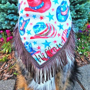 May include: A triangular pet bandana featuring a repeating pattern of red, white, and blue cowboy hats and stars on a light background. The bottom edge is finished with long brown fringe, and the name Willow is embroidered in brown script.