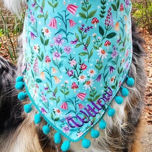 May include: A triangular pet bandana in light blue fabric featuring a colorful botanical print of mushrooms, small flowers, and stars. The bottom edge is trimmed with small turquoise pom-poms, and the name Willow is embroidered in purple.