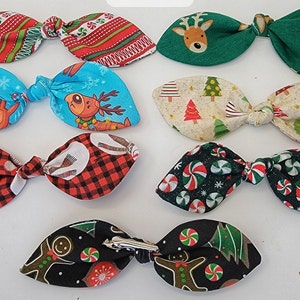 May include: Eight Christmas-themed fabric hair bows with various patterns, including reindeer, candy canes, and Christmas trees. The bows are tied with a knot in the center and have a pointed shape.