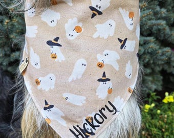 May include: A tan pet bandana featuring a repeating pattern of white ghosts, some wearing black witch hats and holding orange jack-o'-lanterns. The name Hickory is embroidered in black script lettering near the bottom point of the fabric.