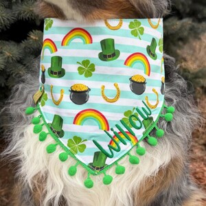 May include: A St. Patrick's Day dog bandana with a repeating pattern of rainbows, green top hats, shamrocks, horseshoes, and pots of gold on a striped background. The bandana is trimmed with green pom-poms and has the name "Willow" embroidered in green.