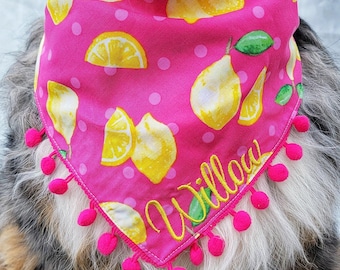 May include: A pink dog bandana with a lemon and pear pattern. The bandana has a repeating pattern of yellow lemons and pears with green leaves and pink polka dots. The word "Willow" is embroidered in yellow. Pink pom-poms trim the edges.