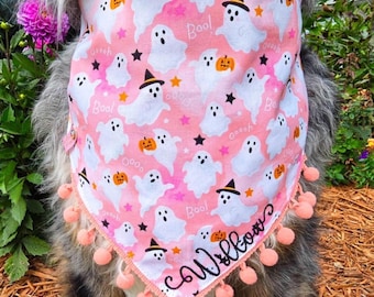 May include: A pink triangular pet bandana featuring a repeating pattern of white ghosts, orange pumpkins, black witch hats, and stars. The fabric includes the text Boo and the name Willow in black script, finished with a light pink pom-pom trim.