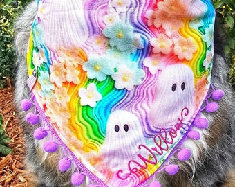 May include: A triangular pet bandana featuring a vibrant rainbow-colored wavy background with white ghost figures and pastel-colored flowers. The fabric is trimmed with small purple pom-poms along the bottom edge and includes the name Willow embroidered in pink cursive script.
