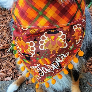 May include: A triangular pet bandana featuring a brown background with a pattern of turkeys wearing pilgrim hats, pumpkins, and acorns. The text "Jasper" is embroidered in white script. The top border is orange plaid, and the bottom edge has orange pom-poms.