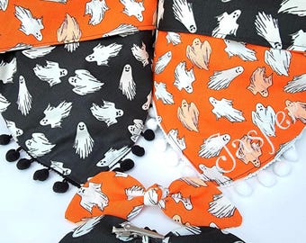 May include: Two triangular pet bandanas featuring a ghost pattern, one with black fabric and black pom-pom trim, the other with orange fabric, white pom-pom trim, and the name Jasper embroidered in white. Two matching hair bows with metal clips.