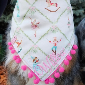 May include: A white triangular pet bandana featuring a green lattice pattern with pink bows and illustrations of nutcrackers, ballerinas, and rocking horses. The name Willow is embroidered in pink script, and the edges are trimmed with pink pom-poms.