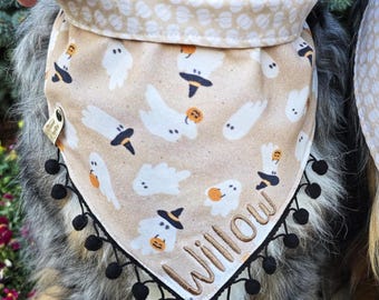 May include: A tan pet bandana featuring a repeating pattern of white ghosts wearing black witch hats and holding orange pumpkins. The bandana is trimmed with small black pom-poms along the bottom edge and features the name Willow embroidered in brown.