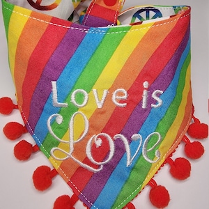 May include: A rainbow-colored bandana with red pom-poms around the edges. The bandana has a white peace sign on the inside and the words "Love is Love" embroidered in white on the front.