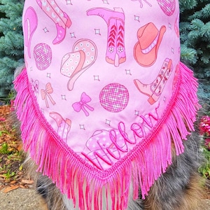 May include: A pink triangular pet bandana featuring a repeating pattern of cowboy boots, hats, disco balls, and bows. The bottom edge is finished with long, bright pink fringe, and the name Willow is embroidered in pink script near the bottom.