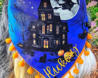May include: A blue triangular pet bandana featuring a haunted house, a full moon with a witch silhouette, bare trees, pumpkins, and a black cat. The name Hickory is embroidered in yellow script, and the edge is trimmed with small orange pom-poms.