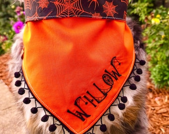 May include: A pet bandana featuring a bright orange main section with the name WILLOW embroidered in black thread, accented by a black top border with a red spiderweb pattern and a black pom-pom trim along the bottom edge of the fabric.