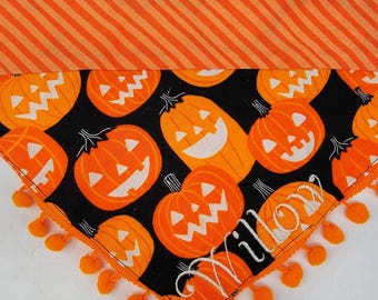 May include: A triangular pet bandana featuring a black background with an orange jack-o'-lantern pattern and an orange striped top border. The name Willow is embroidered in white script, and the bottom edge is trimmed with small orange pom-poms.