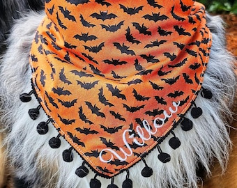 May include: A triangular pet bandana featuring an orange fabric with a repeating black bat pattern. The bottom edge is trimmed with small black pom-poms. The name Willow is embroidered in white cursive script across the lower right section.