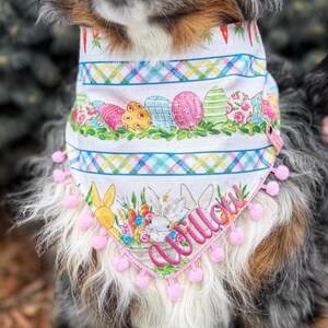 May include: A white dog bandana with an Easter theme. The bandana features colorful Easter eggs, bunnies, and carrots. The bottom edge is trimmed with pink pom-poms, and the name "Willow" is embroidered in pink script.
